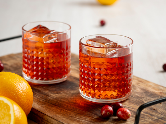Zwei Gläser mit rotem Aperitif auf Eis, serviert mit Orangen und Cranberries auf einem rustikalen Holztablett.