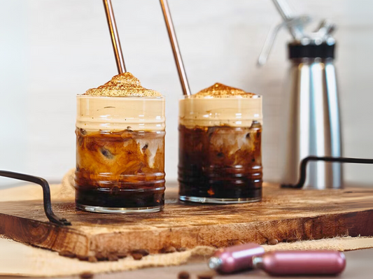 Zwei Iced Coffees mit cremiger Espuma-Krone und Strohhalm, verfeinert mit Kakao – zubereitet mit dem iSi Gourmet Whip, serviert auf rustikalem Holzbrett.